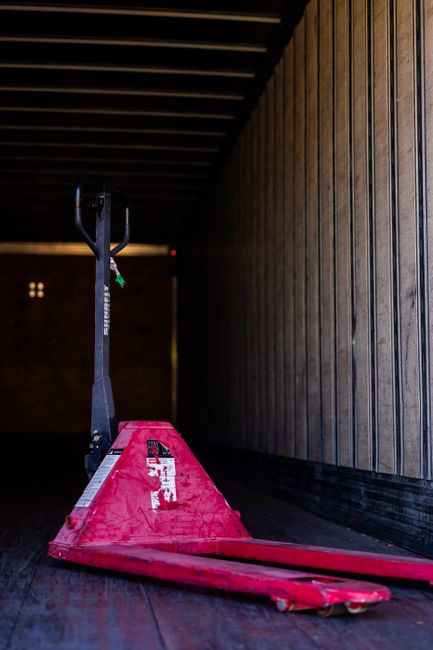 Photograph showing a home relocation scene with a furniture removal process in progress. Inside a residential property, a large wooden wardrobe and several cardboard boxes, some wrapped in plastic and