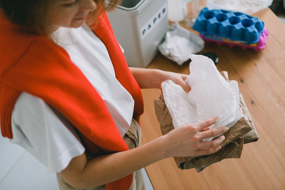 A person in a white shirt with a red and orange knitted cardigan draped over their shoulders is holding a piece of crumpled white tissue paper and a brown paper bag, preparing packing materials for ho