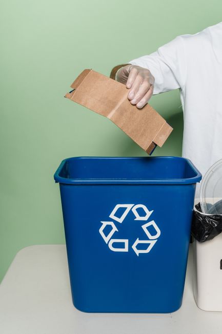 A person in a white shirt with a red and orange knitted cardigan draped over their shoulders is holding a piece of crumpled white tissue paper and a brown paper bag, preparing packing materials for ho