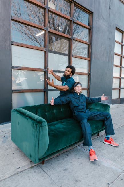 Two men are positioned outdoors in front of a modern building with large glass windows divided by wooden frames; one man stands behind a dark green upholstered sofa, using both hands to steady it whil