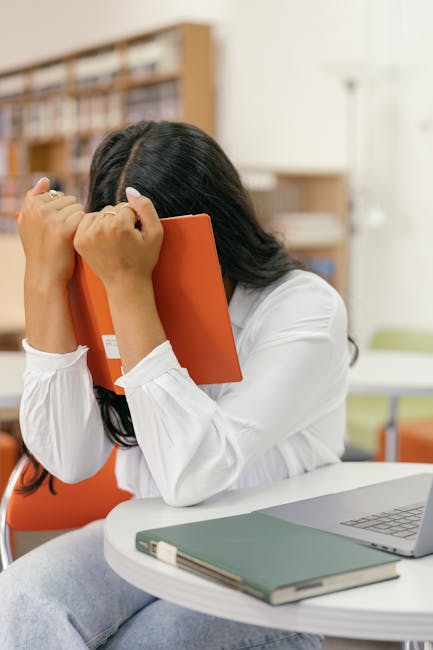 A person sitting on a beige fabric armchair while working on a silver laptop, which displays several colorful stickers with programming-related icons and text such as 'PHP' and '<>'. The person is wea