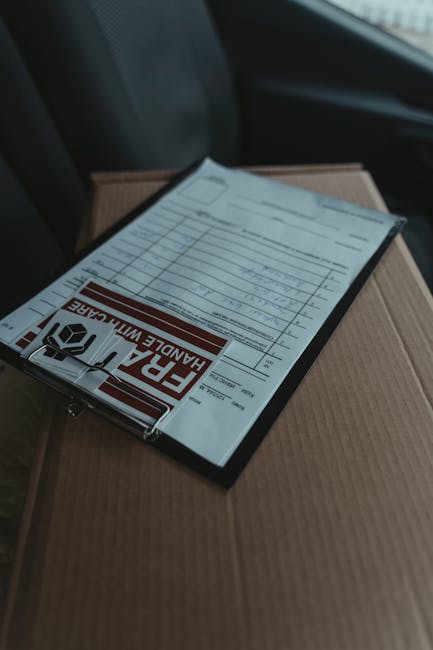 A close-up view of a clear plastic clipboard with a handle, resting on top of a brown cardboard box inside a vehicle. The clipboard holds a printed handle-with-care warning label in red and white, alo