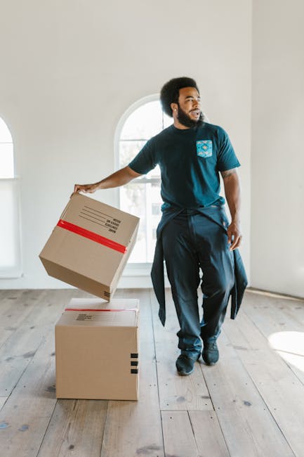 A woman with curly black hair and a bright smile is seated on a chair inside a room, surrounded by stacked cardboard moving boxes in various sizes, some sealed with red or black tape. She is wearing a